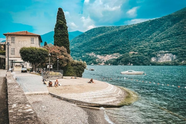Un passeig al costat del llac amb una banca de pedra, un arbre alt i un edifici amb teulada vermella.