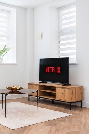 Una sala moderna con un mueble bajo para entretenimiento de madera, un televisor de pantalla plana mostrando el logotipo de Netflix, una mesita con un cuenco decorativo y una planta en maceta.