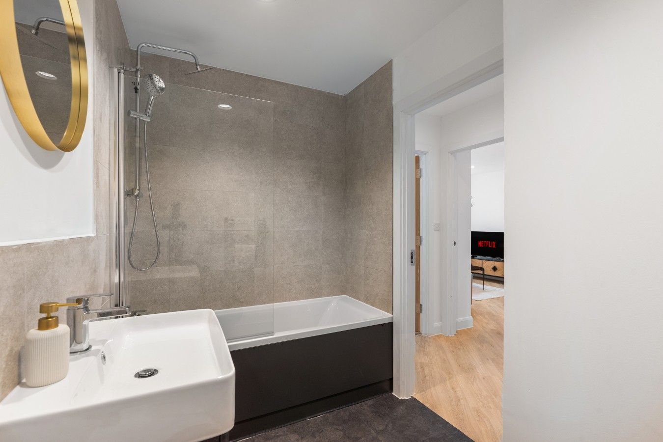 Un baño moderno con bañera, ducha y lavamanos. Se observa un espejo con marco dorado y un dispensador de papel higiénico.