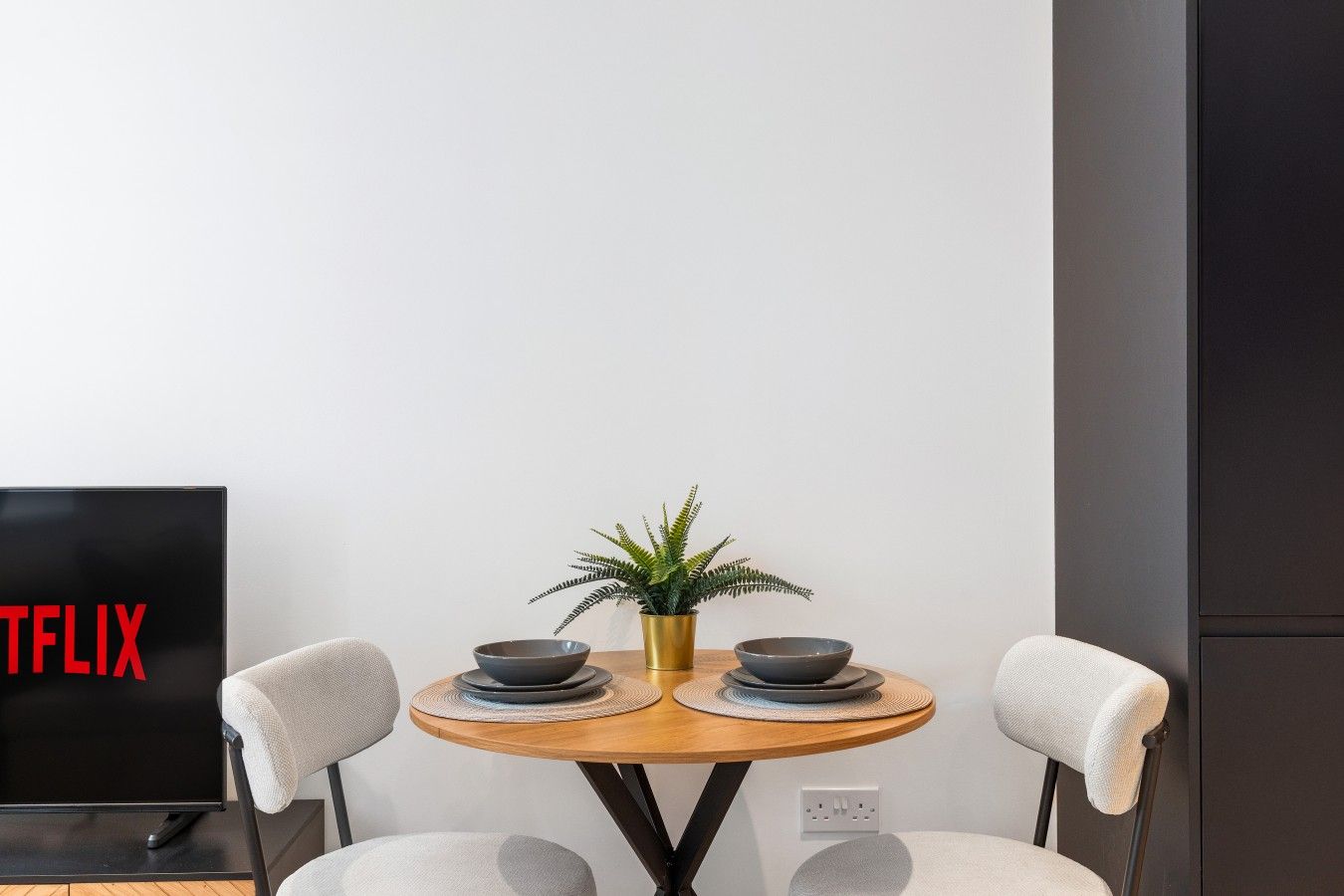 Un comedor moderno con una mesa redonda, dos sillas, una planta en maceta y un televisor que muestra el logotipo de Netflix.