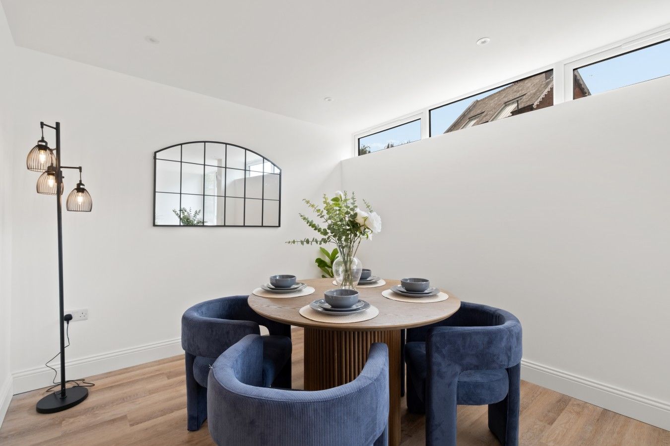 Uma sala de jantar com mesa redonda, cadeiras estofadas em azul, um vaso com flores e uma luminária pendente moderna.