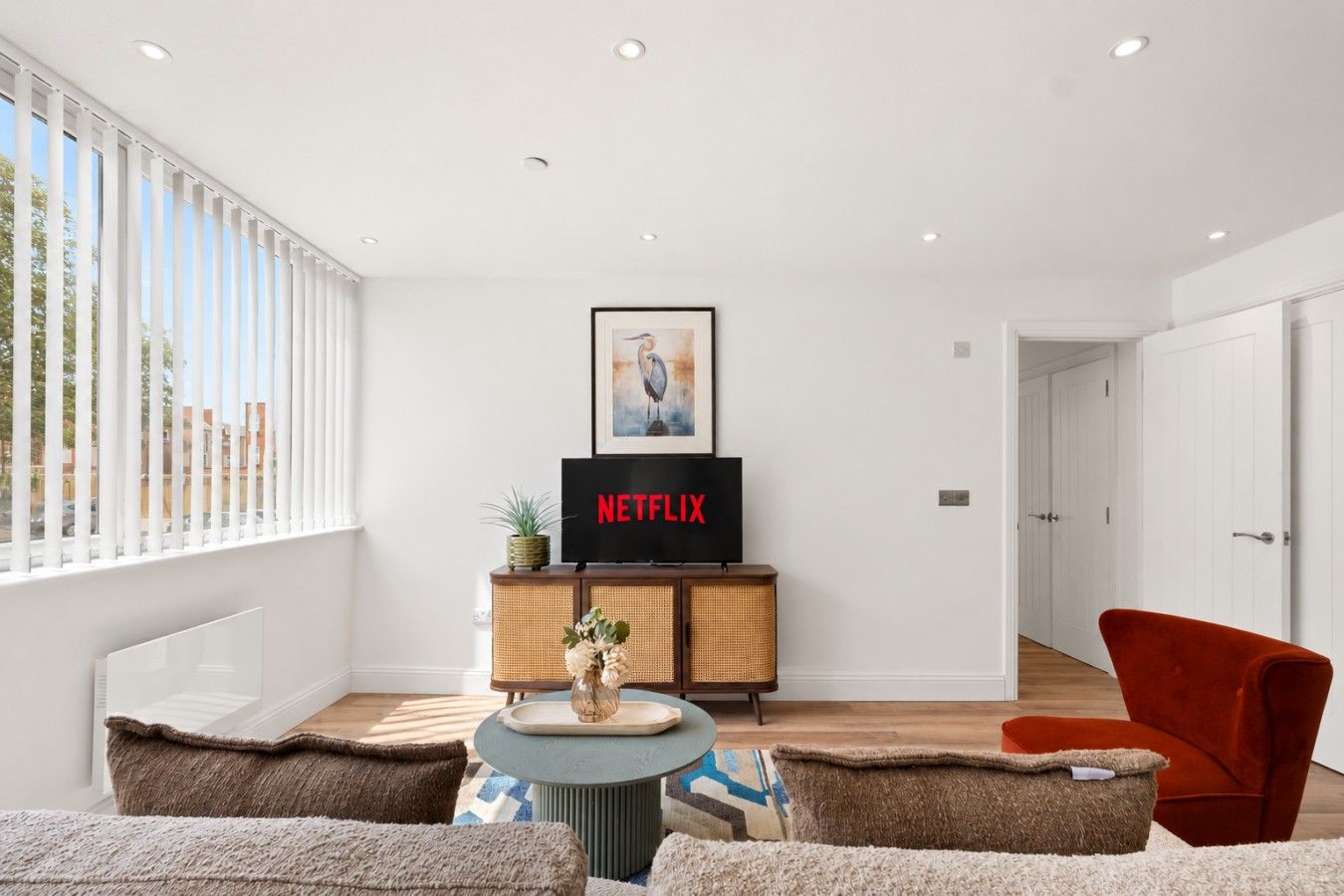 Sala de estar moderna com televisão de tela plana exibindo 'NETFLIX', uma obra de arte emoldurada acima da TV, um aparador de vime, uma mesa redonda pequena com um vaso e uma poltrona vermelha.
