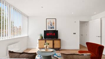 Sala de estar moderna com televisão de tela plana exibindo 'NETFLIX', uma obra de arte emoldurada acima da TV, um aparador de vime, uma mesa redonda pequena com um vaso e uma poltrona vermelha.