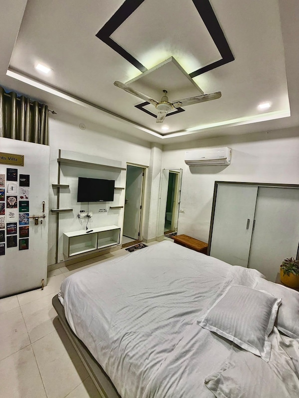 A hotel room with a bed, a TV mounted on the wall, a wardrobe, and a ceiling fan. - Hotel 1947 (Pushkar)
