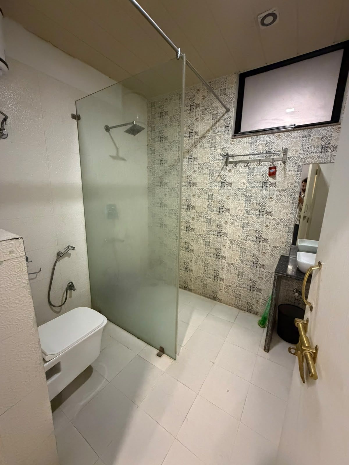 Un baño con ducha de vidrio, inodoro blanco, lavamanos con espejo y televisor montado en la pared.