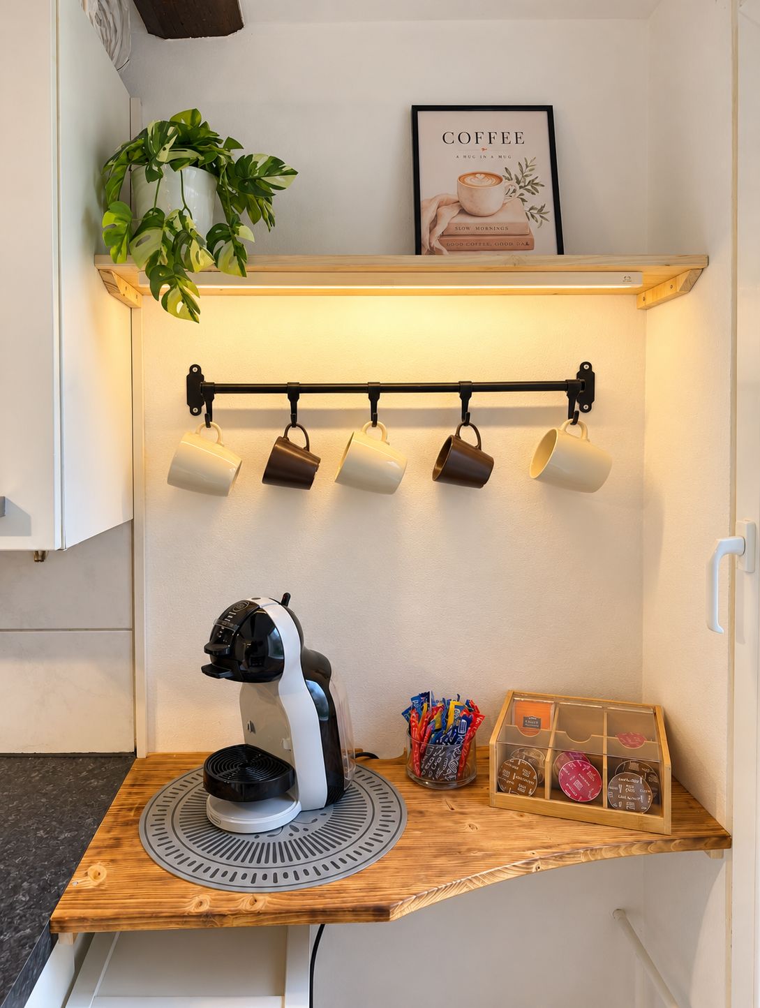 Een keukenhoek met een koffieapparaat, een ophangrek voor mokken, een potplant en een kunstwerk met koffiethema.