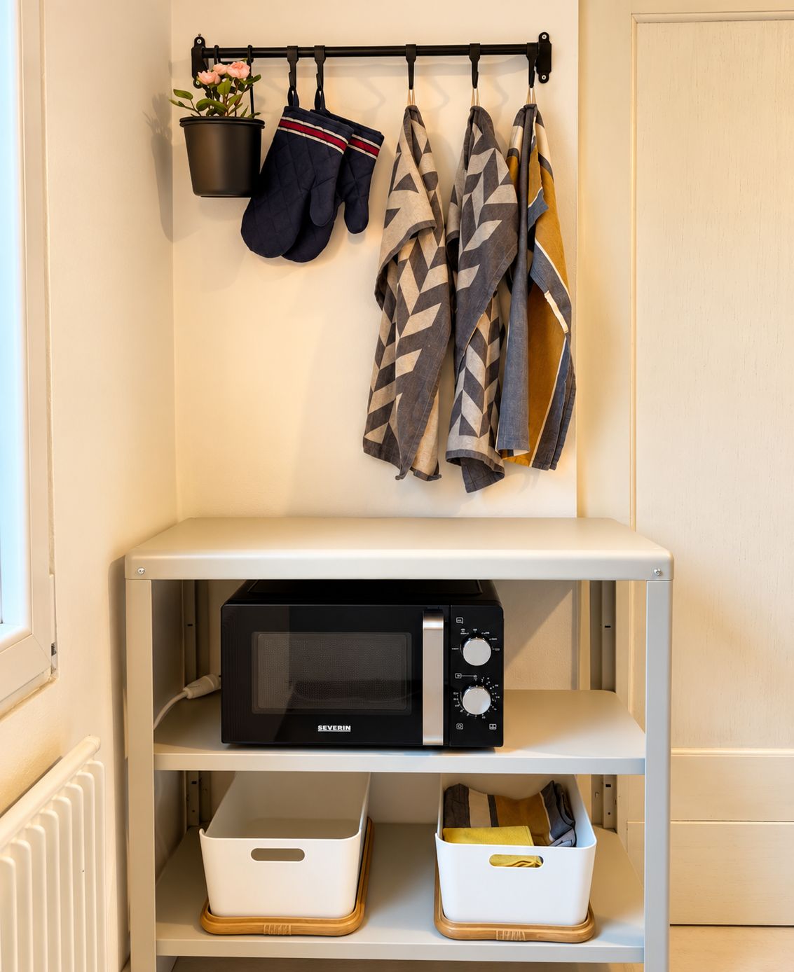 Un coin cuisine avec un four à micro-ondes posé sur une étagère blanche, un porte-ustensiles suspendu avec des gants et une veste à motifs, et deux bacs de rangement blancs dotés de poignées.