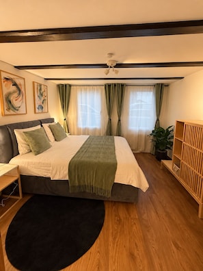 Une chambre à coucher avec un lit, des tables de chevet, un tapis noir, un plancher en bois, un ventilateur de plafond et une plante.