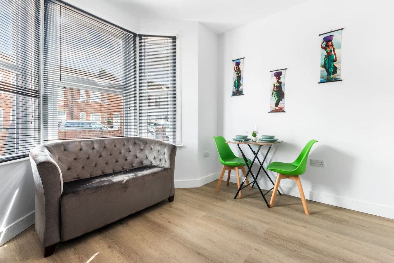 A room with a grey sofa, a small table with green chairs, and three framed pictures on the wall.