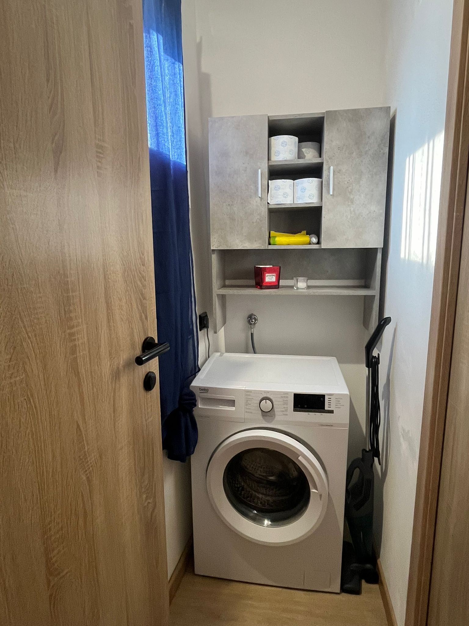 A compact laundry room with a front-loading washing machine, a shelf with toiletries, and a door with a blue curtain.
