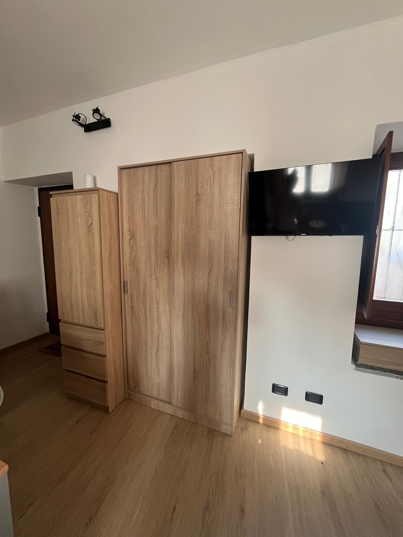 A room with a wooden wardrobe, a white wall, and a window.