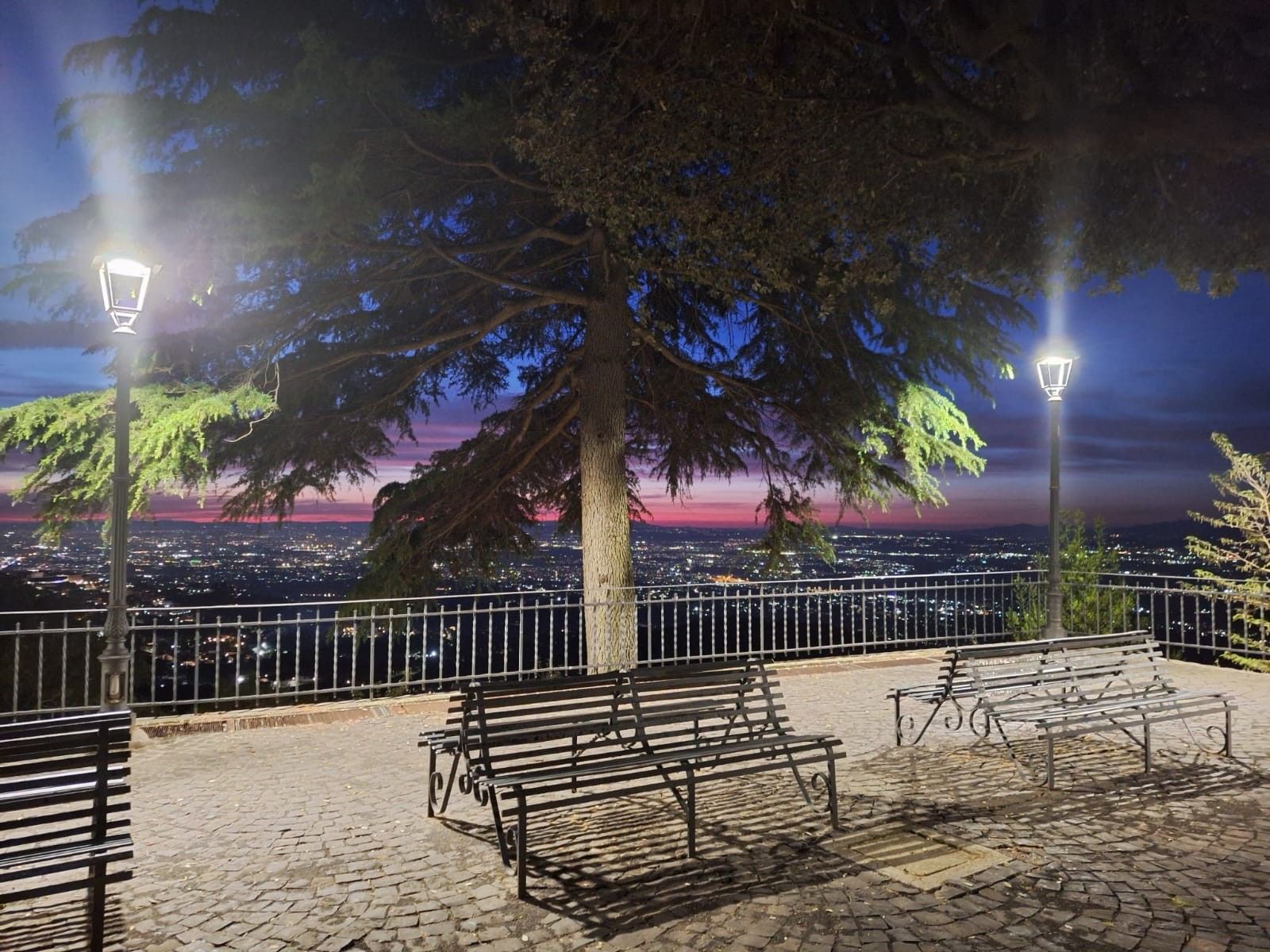 Un área de estar al aire libre con bancos y faroles, con vista a un paisaje urbano al atardecer.