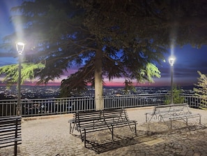 Un área de estar al aire libre con bancos y faroles, con vista a un paisaje urbano al atardecer.
