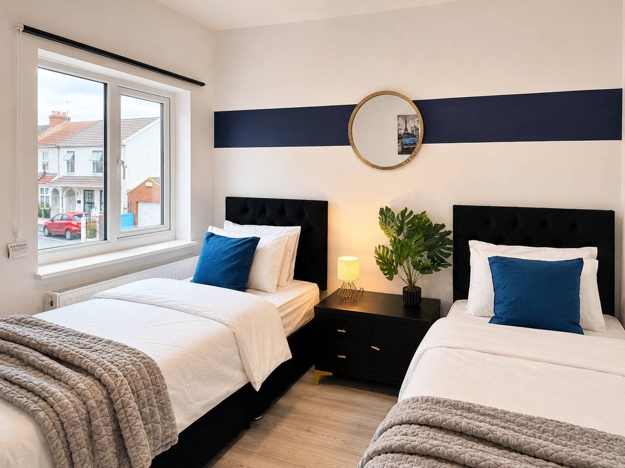 A hotel room with two beds, a window with a view of the street, a bedside table with a lamp, and a wall-mounted mirror.