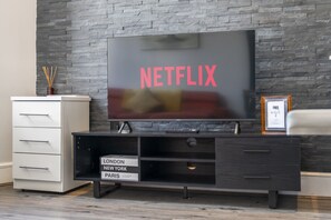 Ein modernes Wohnzimmer mit einem Flachbildfernseher, der 'NETFLIX' in roten Buchstaben anzeigt, einer niedrigen Unterhaltungsanlage aus dunklem Holz mit Schubladen und einem gerahmten Bild an der Wand.
