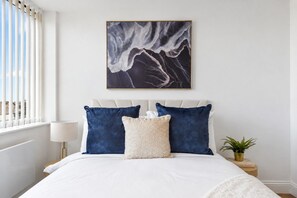 Quarto com cama, almofadas, uma obra de arte na parede, abajur e uma pequena planta.
