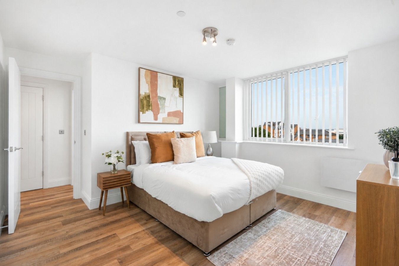 Quarto com uma cama grande, mesa de cabeceira com abajur, janela com persianas e porta.