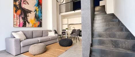 A modern living room with a grey sofa, a colorful abstract painting, a staircase, and a dining area in the background.
