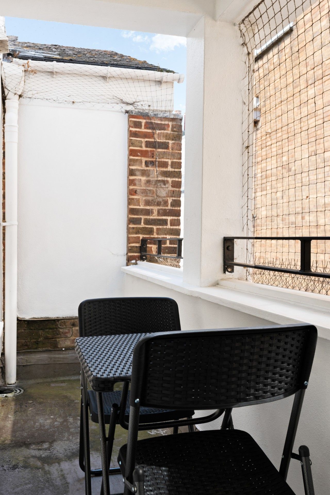 A balcony with two chairs and a table, a white wall, a brick wall, and a window with a metal grille.
