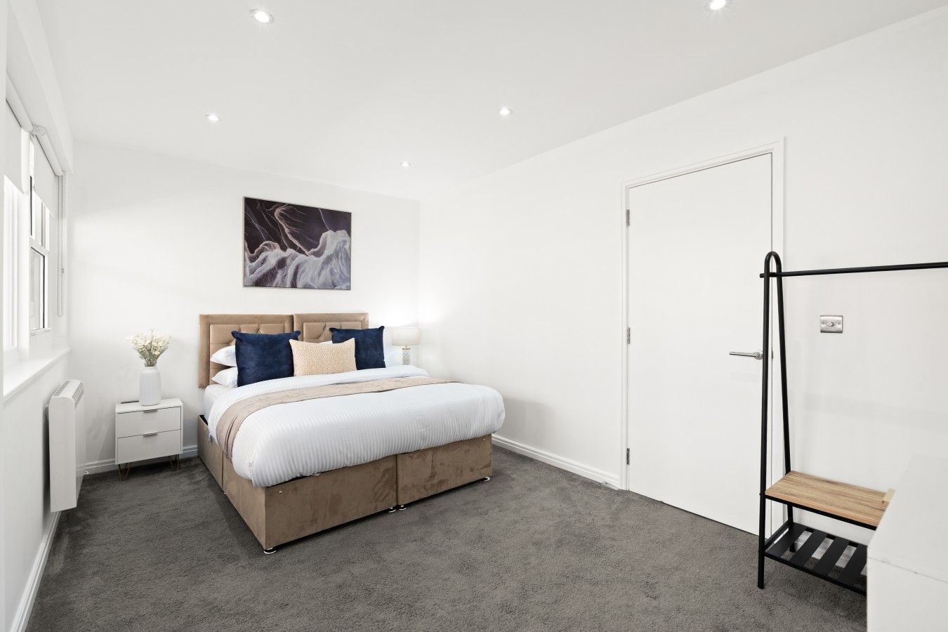 A modern bedroom with a bed, bedside tables, a nightstand, a wall-mounted artwork, a door, and a small wooden bench.