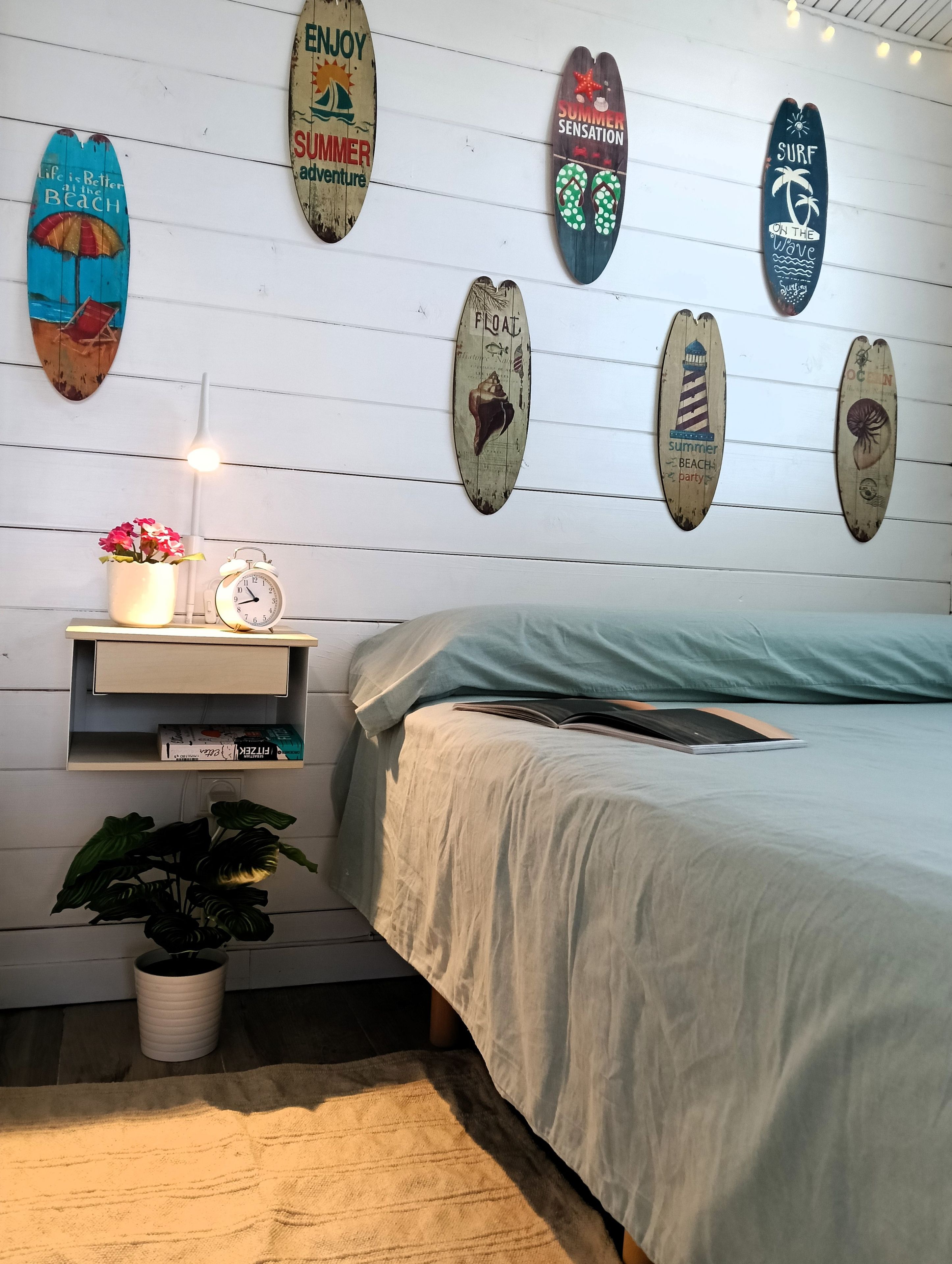 Een slaapkamer met een bed, een nachtkastje met een lamp, een plank met boeken, en surfplanken aan de muur.