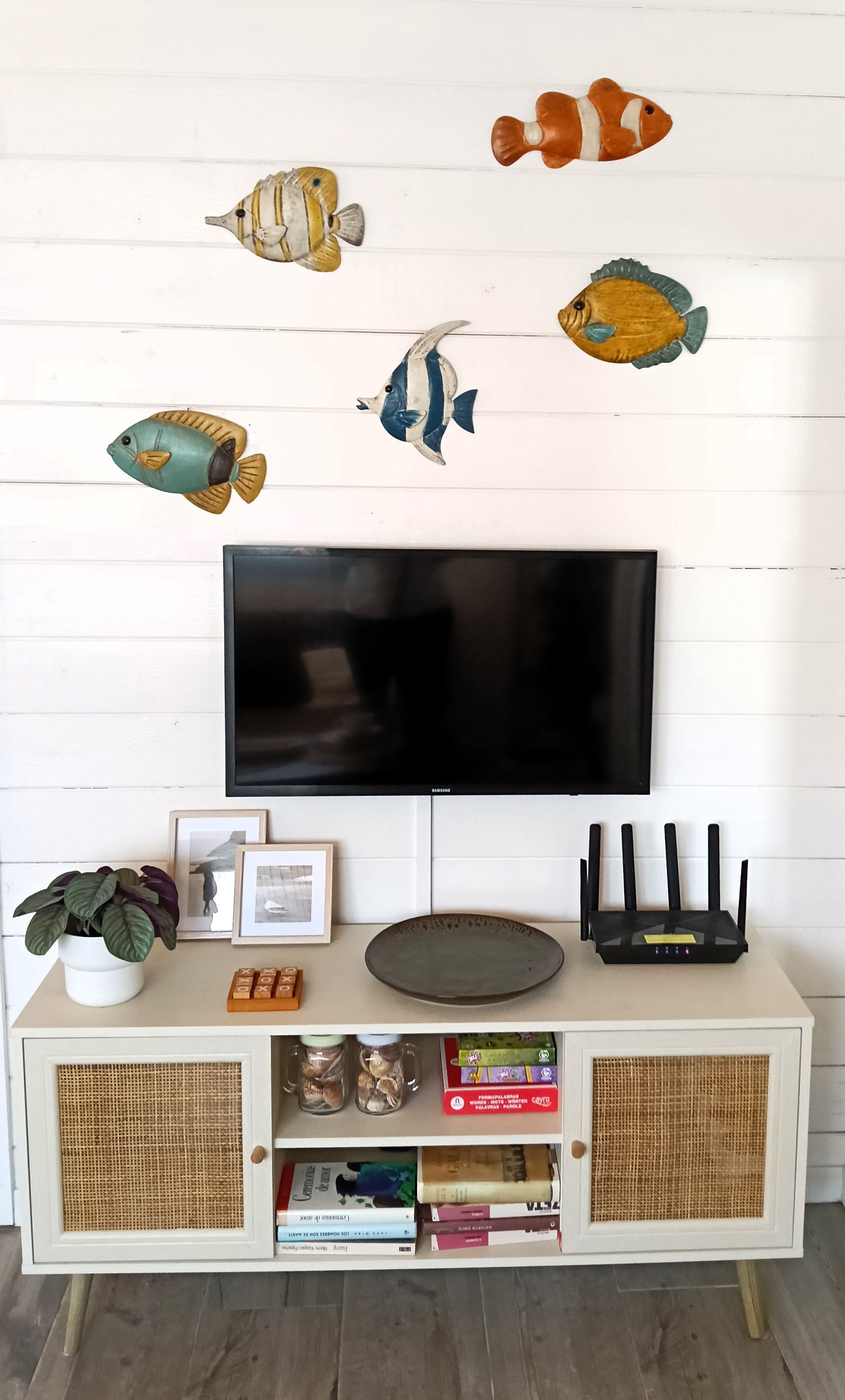 Een flatscreen-tv bevestigd aan een witte muur met decoratieve visstickers, een potplant, ingelijste foto's, een router en een boekenplank met boeken en decoratieve objecten.
