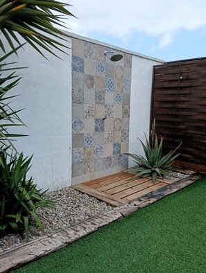 Een betegelde douche ruimte met een houten deur, omgeven door een tuin met kunstgras, kiezelstenen en planten.