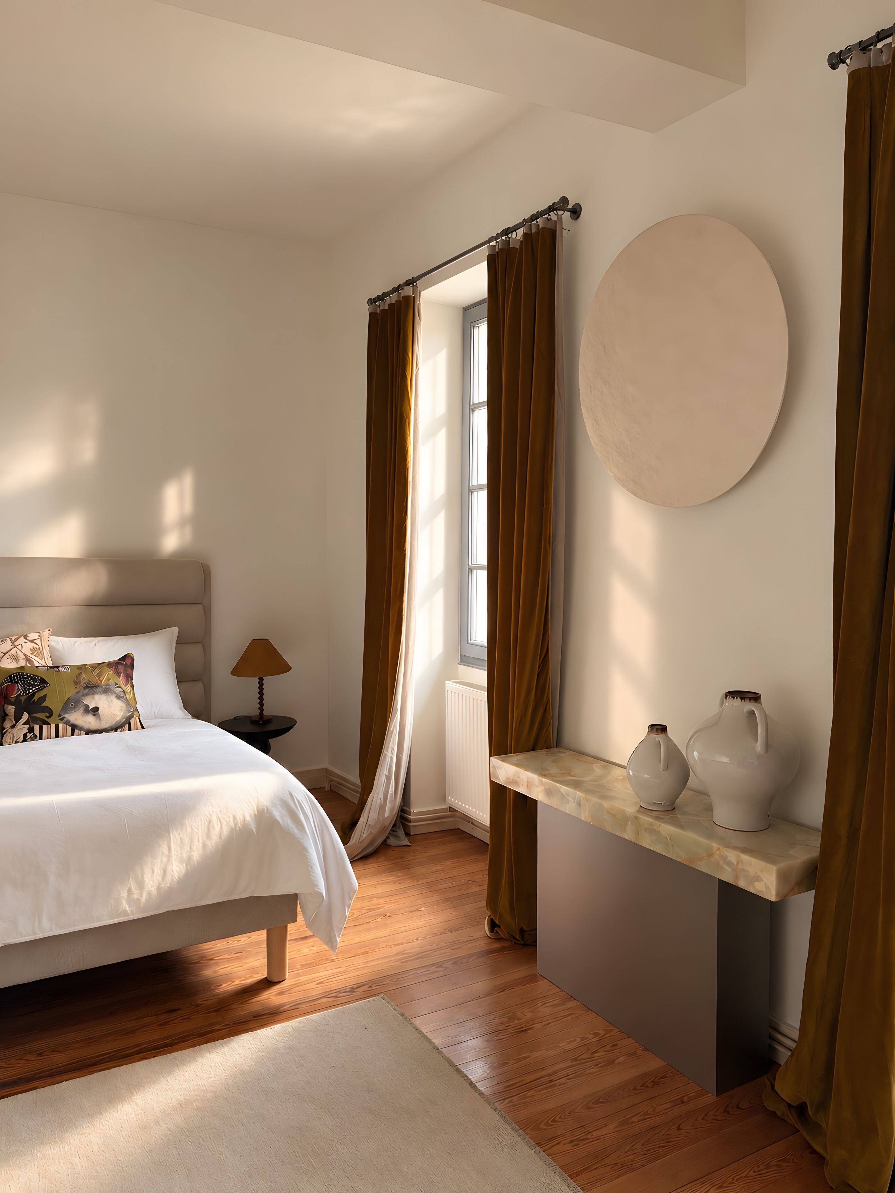 Une chambre à coucher avec un lit, une table de chevet ornée de deux vases, une fenêtre avec des rideaux et un radiateur.
