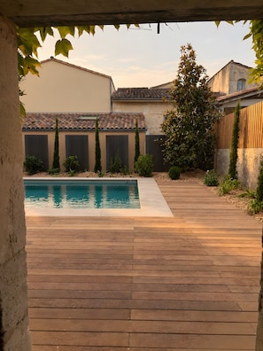 Eine Holzterrasse führt zu einem Schwimmbecken mit einer Steinmauer und einem Gebäude im Hintergrund.