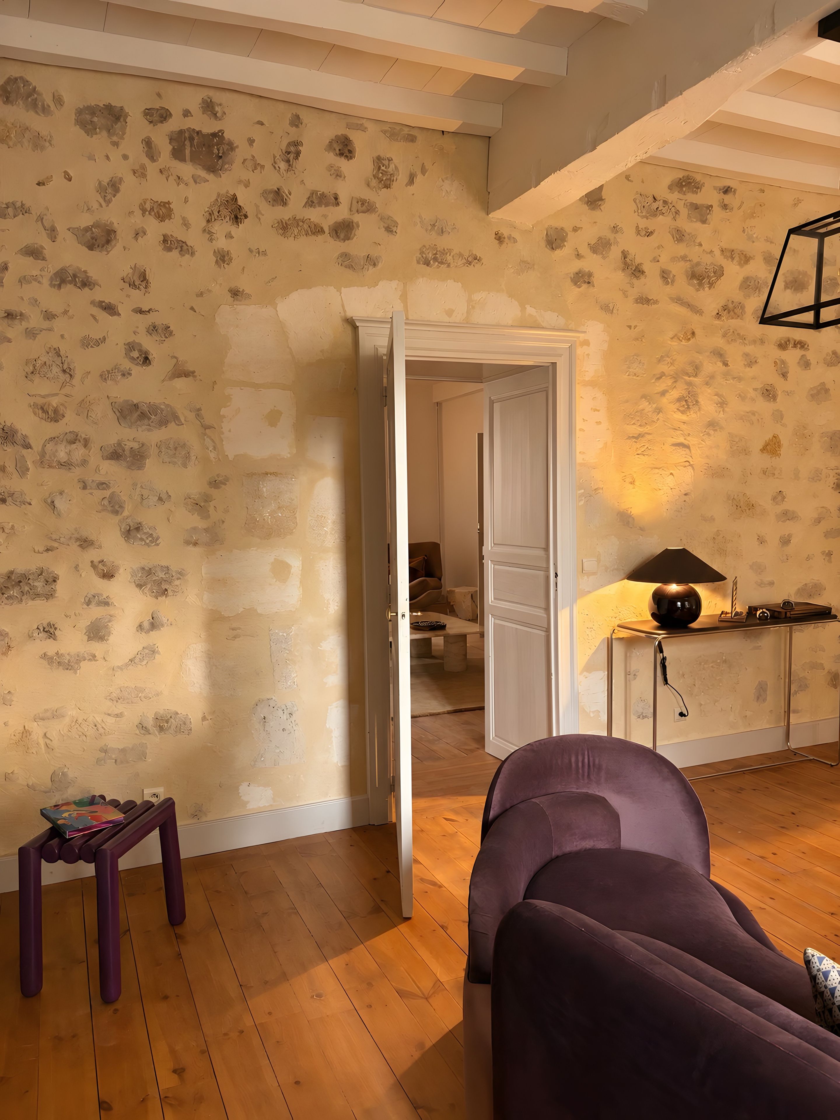 Une pièce avec un plancher en bois, un canapé violet, une petite table avec des livres et une porte qui donne sur une autre pièce.