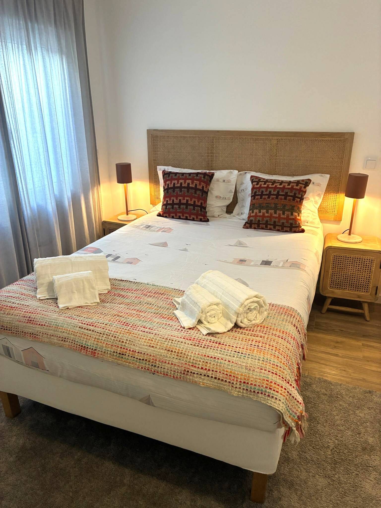 Een netjes opgemaakt bed met een bedsprei met dessin, twee kussens en een set handdoeken ernaast. Aan beide zijden van het bed staan nachtkastjes met een lamp.