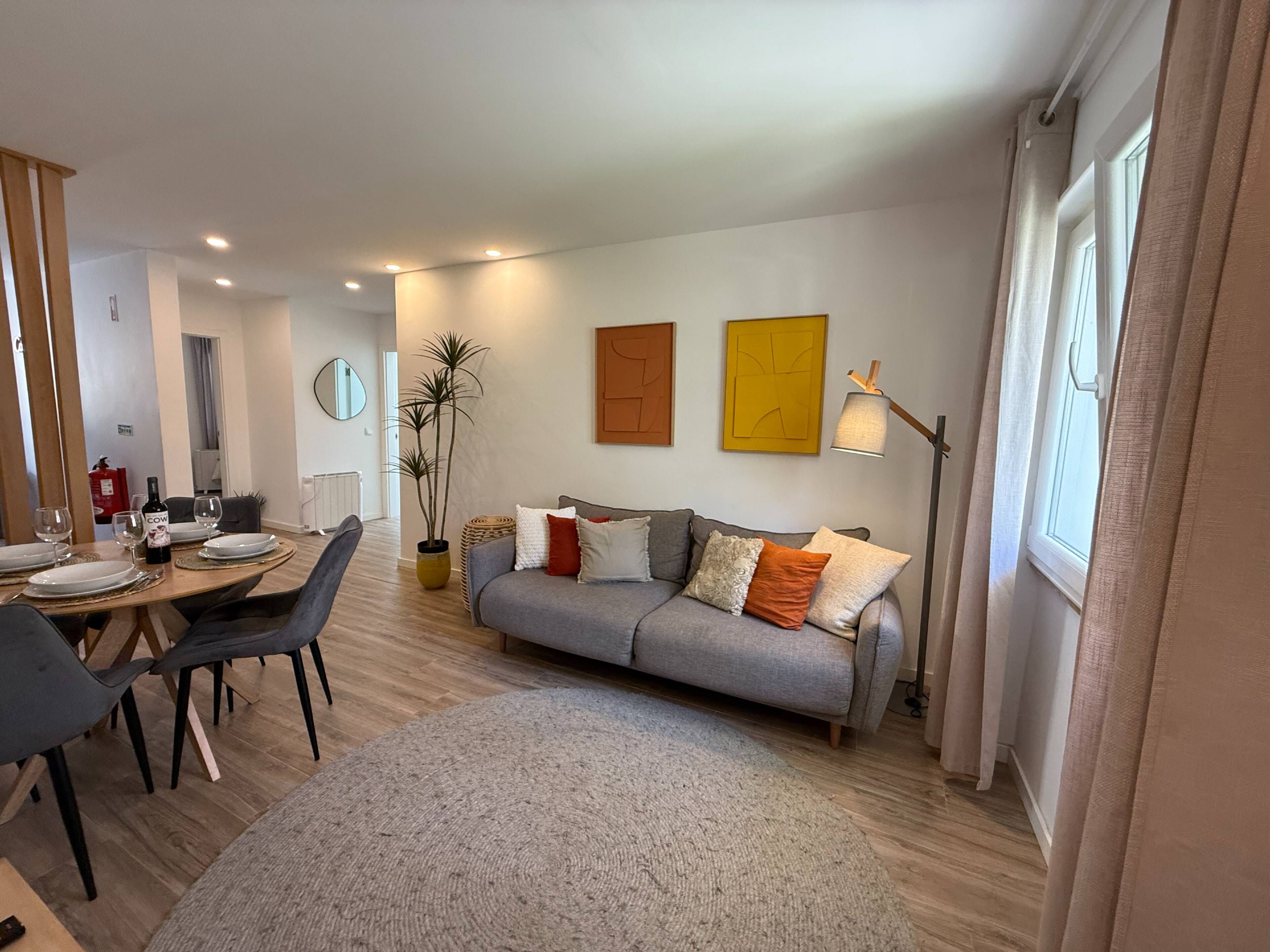 Een moderne woonkamer met een grijze bank, een eettafel met stoelen, een vloerlamp en een potplant.