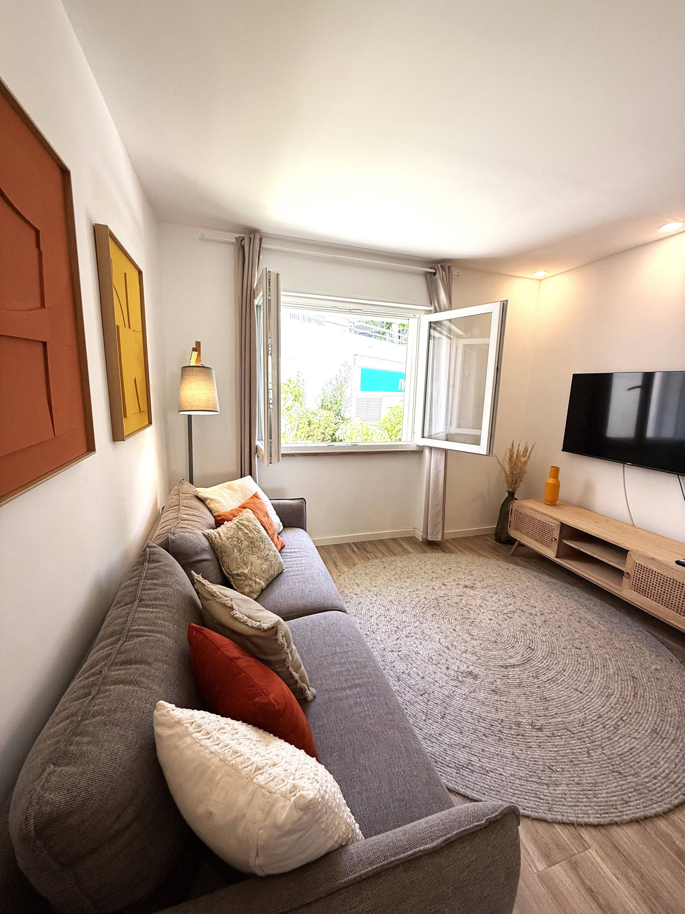 Una sala de estar moderna con un sofá gris, un televisor de pantalla plana, una ventana con cortinas y una lámpara.