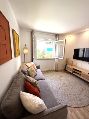 Ein modernes Wohnzimmer mit einem grauen Sofa, einem Flachbildfernseher, einem Fenster mit Vorhängen und einer Lampe.