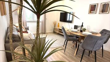 A modern living room with a dining area, featuring a large plant, a sofa, a TV, and a wooden floor.