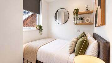 A bedroom with a bed, a bedside lamp, a round mirror, a window with blinds, and a shelf with plants and a decorative item.