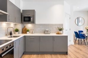 A modern kitchen with grey cabinetry, a microwave, a toaster, and a dining area with blue chairs. - Coulsdon Central Stay | 2 Bedrooms | Sleeps 5 (Coulsdon)