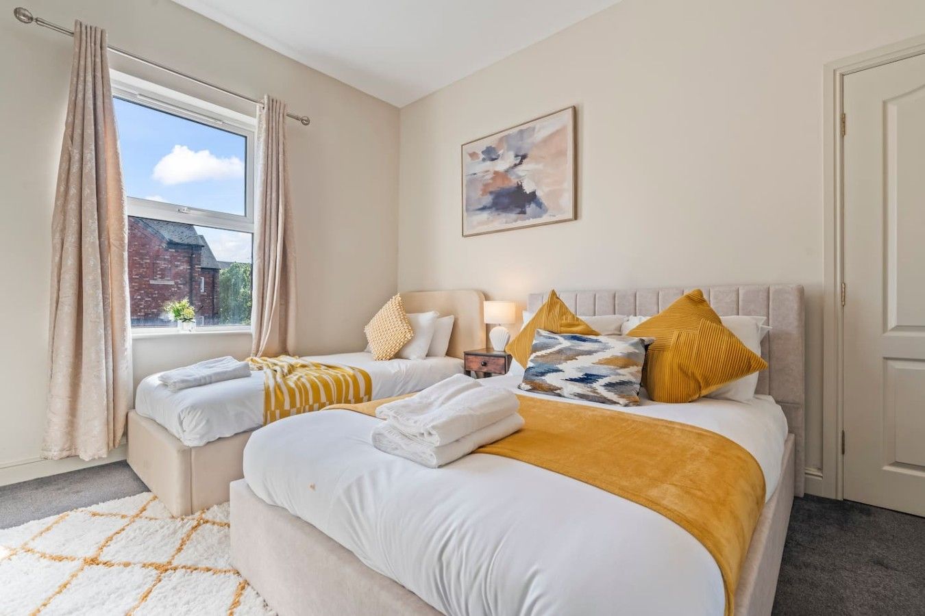 A hotel room with two beds, a window with curtains, a painting on the wall, and a door.