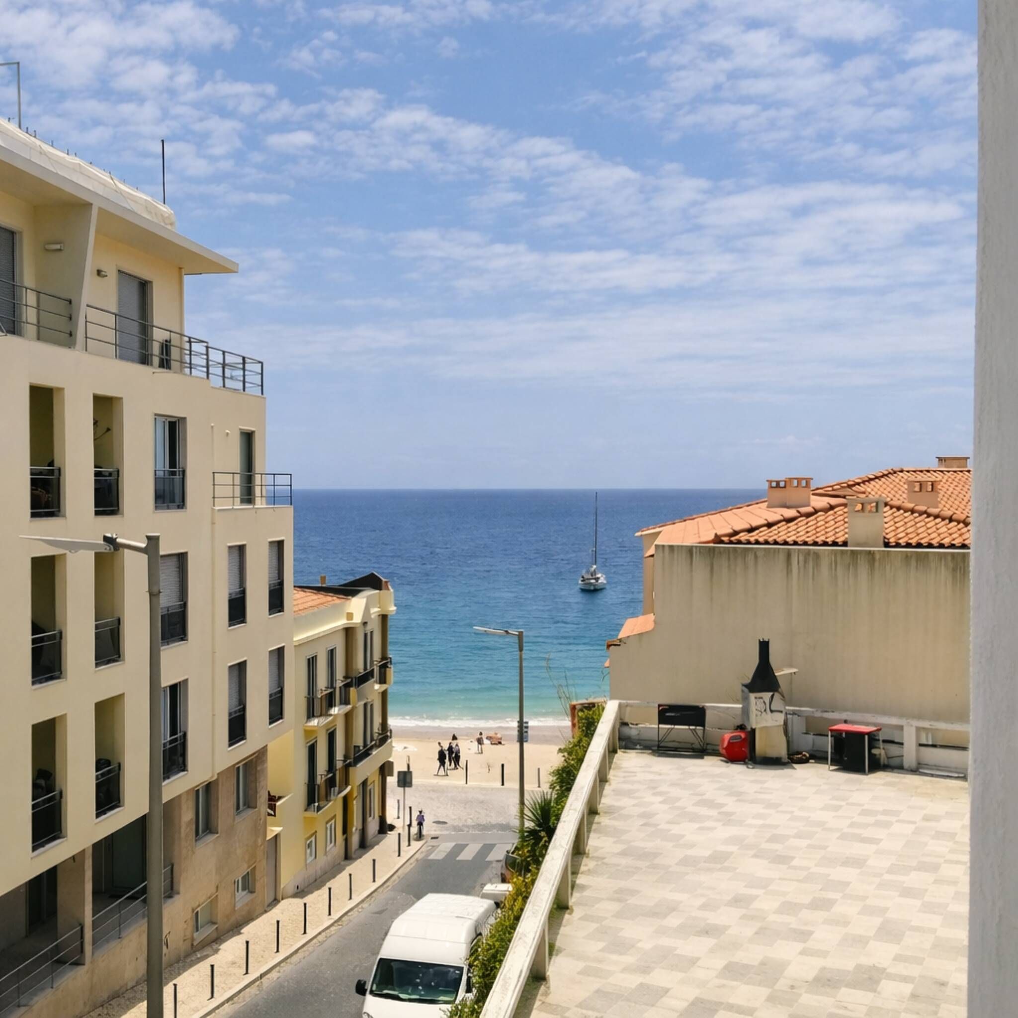 Un bâtiment situé sur la côte, doté de balcons donnant sur la mer, sous un ciel dégagé, avec un voilier visible au loin.