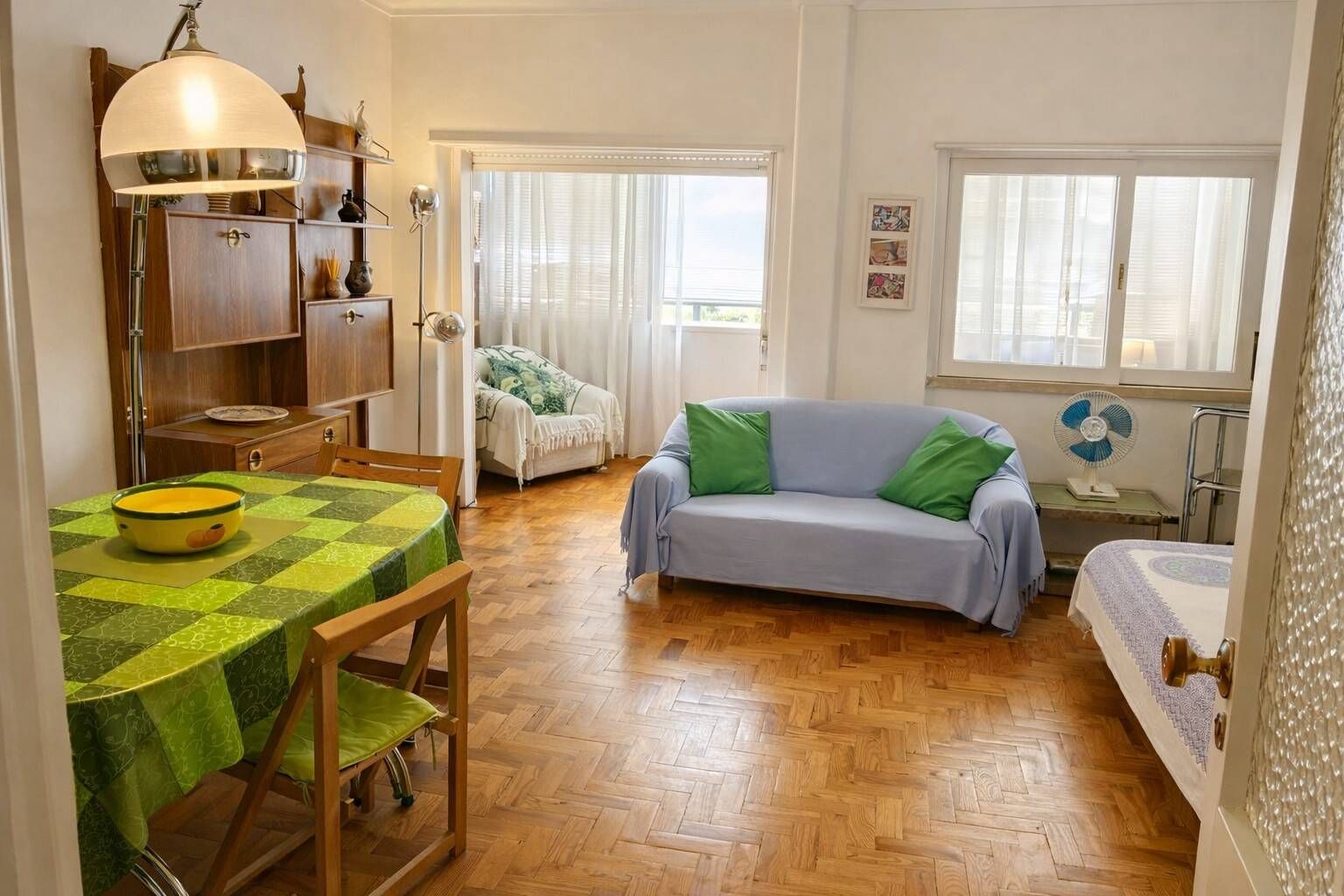 Ein gemütliches Wohnzimmer mit einer Couch, einem Esstisch mit einer Schüssel, einem Bücherregal und einem Ventilator.