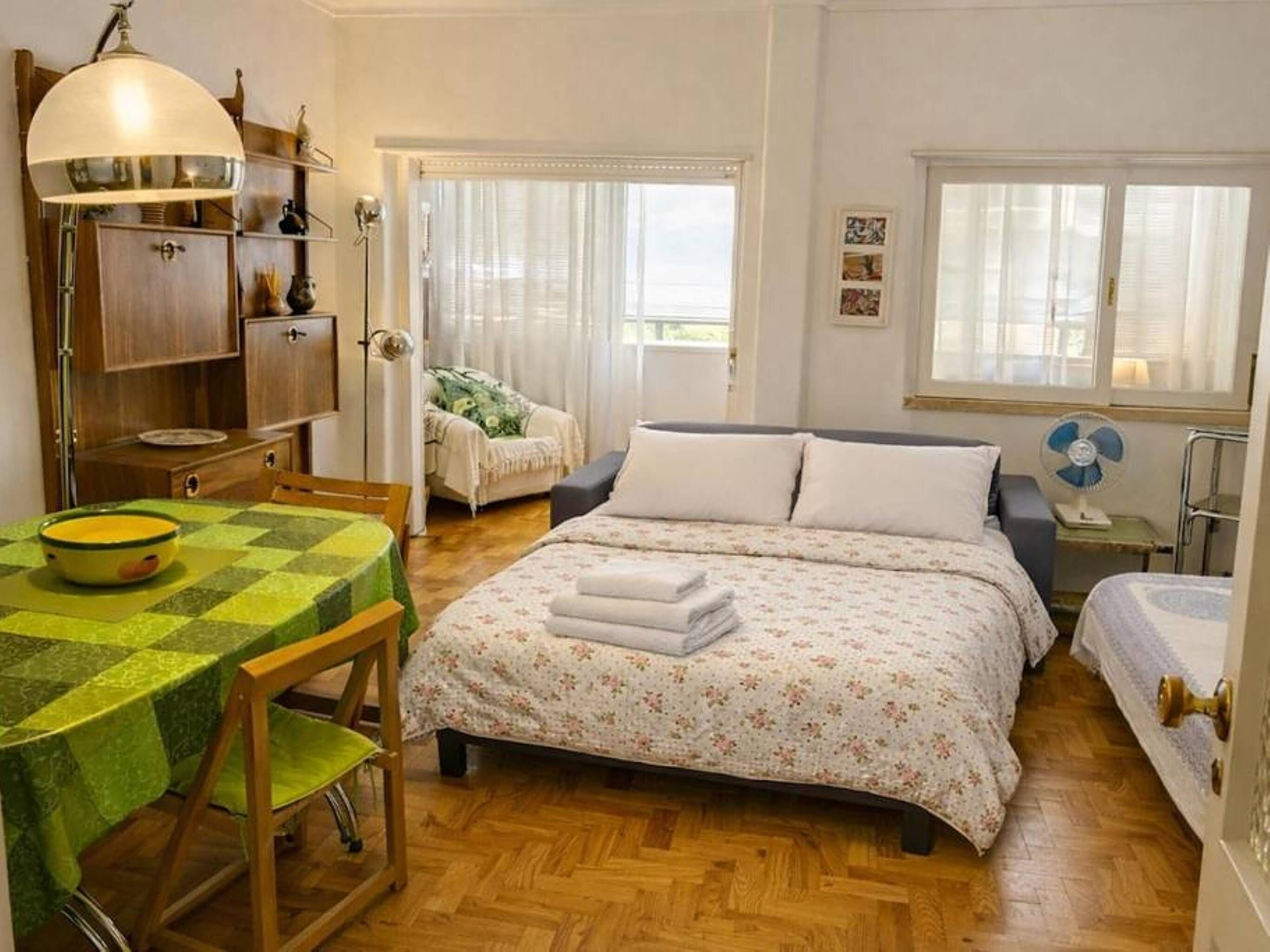 Una camera da letto con un letto, un tavolo con una ciotola, una sedia, un comodino con una ventola e una finestra con tende.