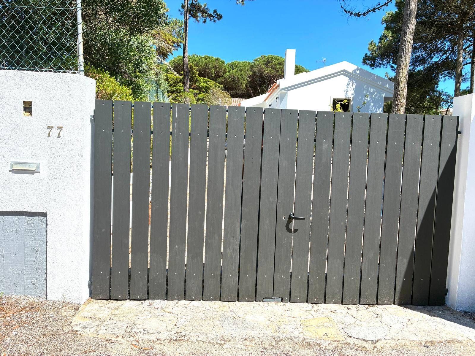 A gated entrance with a number '77' on a white wall, a metal fence, and a clear sky.