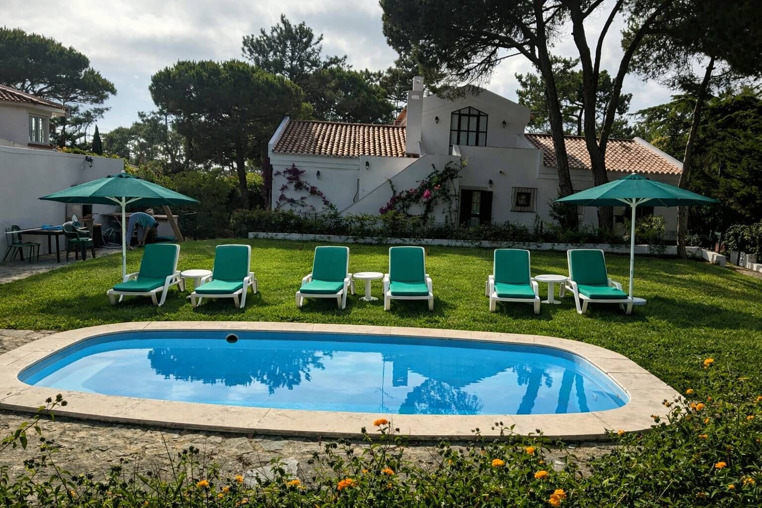 A swimming pool with green lounge chairs and umbrellas, surrounded by a well-maintained lawn and a building with a tiled roof.