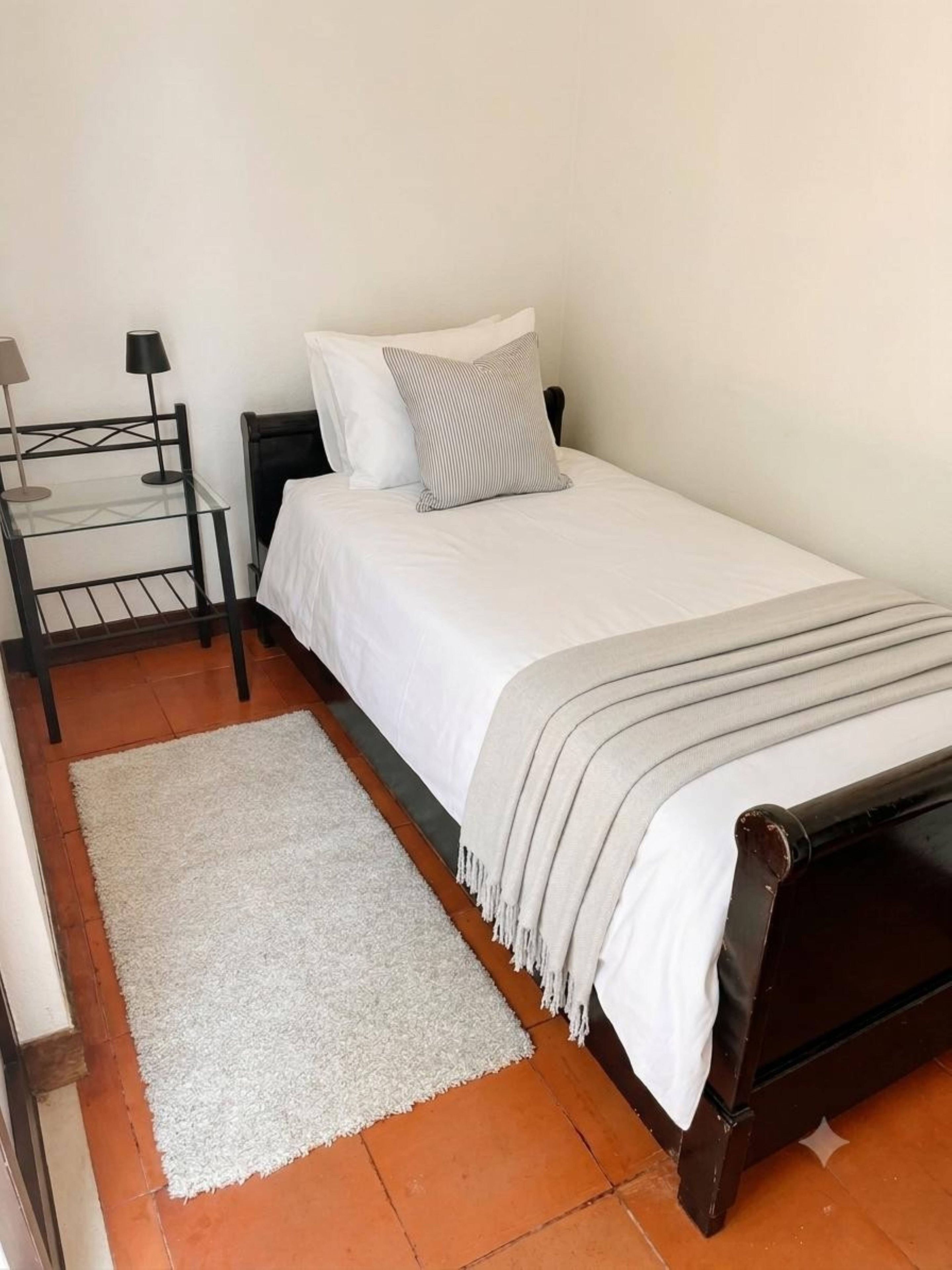 A single bed with a white mattress, a grey and white striped pillow, and a grey blanket. There is a small table with a lamp beside the bed.
