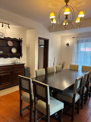 A dining area with a large table and chairs, a chandelier, and a wall-mounted shelf with plates. - Pinewood Beach Villa- Colares (Sintra)