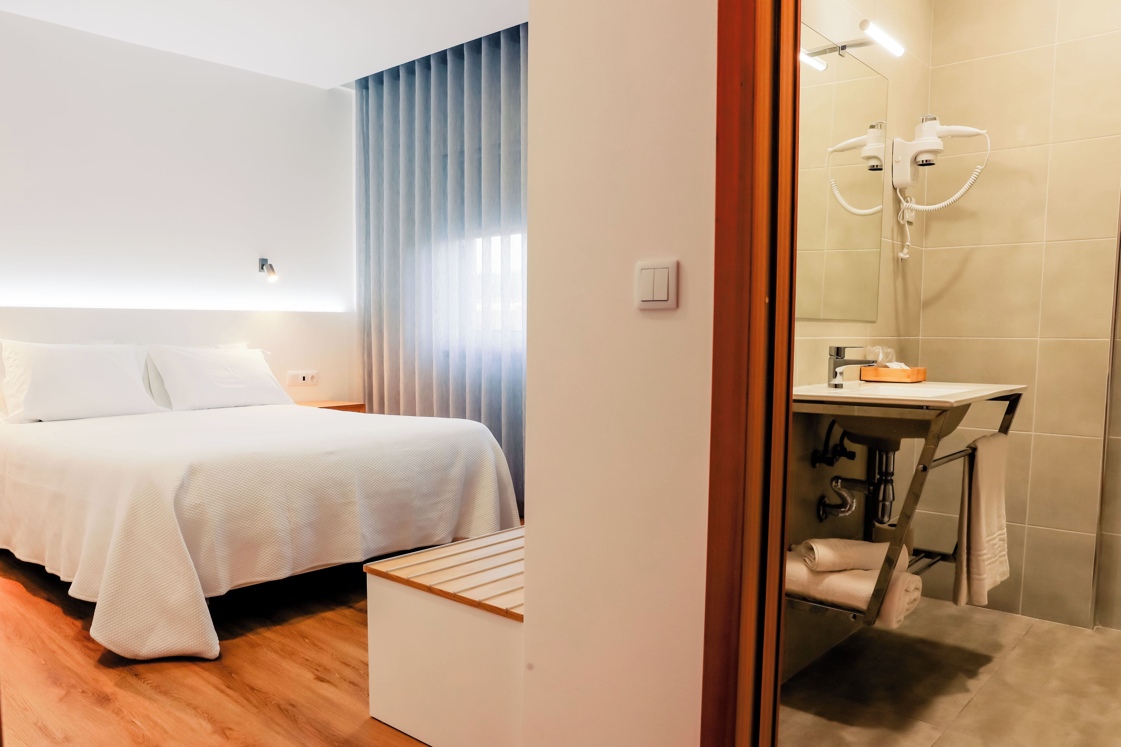 Habitación de hotel moderna con una cama bien hecha, un baño con ducha y lavamanos, y una pequeña mesita de noche de madera.