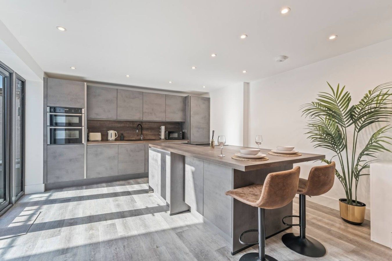 A modern kitchen with a central island, bar stools, and a large potted plant.