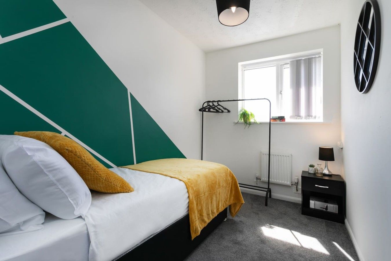 A bedroom with a bed, a radiator, a window with curtains, a plant, and a bedside table with a lamp.