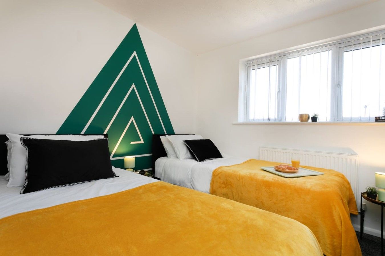 Ein Hotelzimmer mit zwei Betten, einer großen geometrischen Wanddekoration, einem Fenster mit Jalousien und einem kleinen Tischchen mit Tablett.