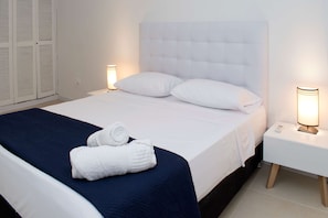 Una cama bien hecha con sábanas blancas, una toalla doblada y dos lámparas de noche.